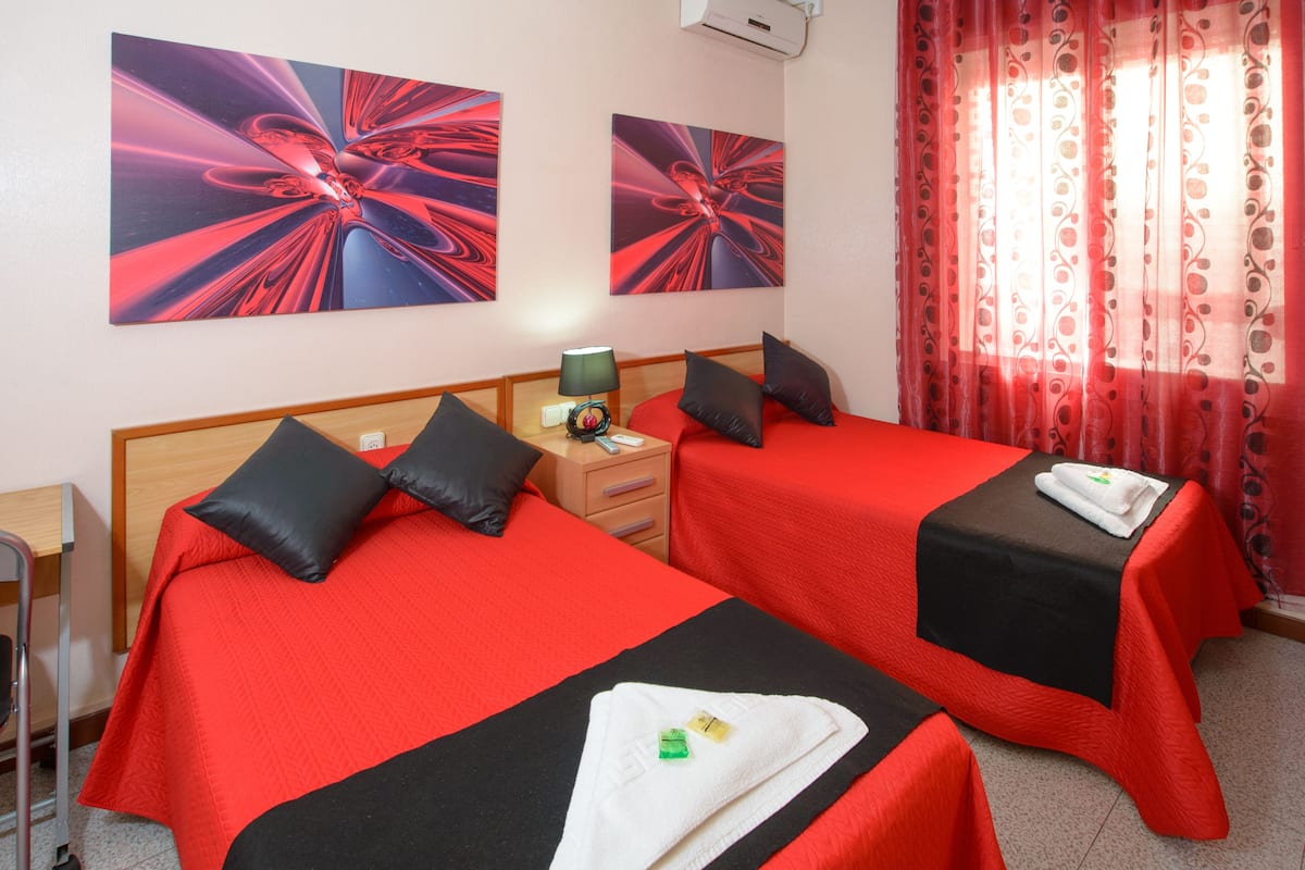 A hotel room with two beds, each with red bedding and black pillows, a bedside table with a lamp, and artwork on the walls.