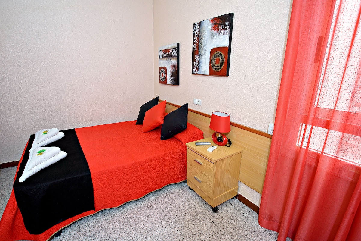 A hotel room with a bed, a wooden nightstand, a red lamp, and artwork on the walls.