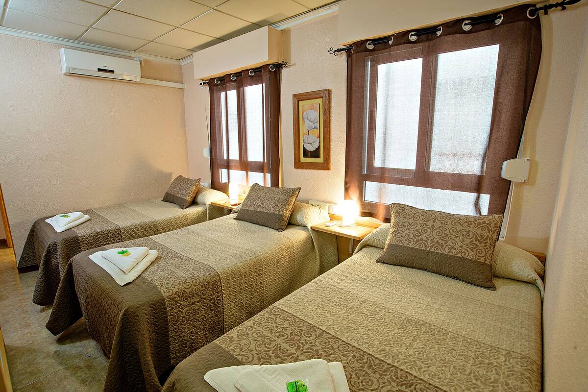 A hotel room with two beds, each with a patterned bedspread and pillows, a bedside table with a lamp, and an air conditioning unit on the wall.
