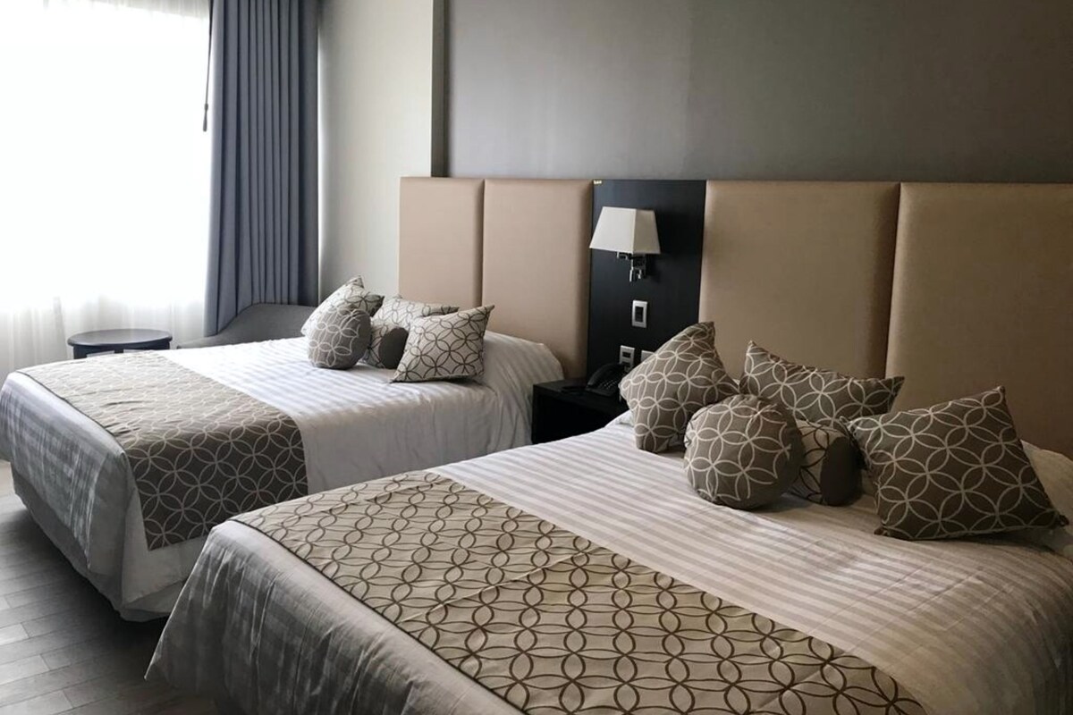Habitación de hotel con dos camas, cada una con ropa de cama y almohadas estampadas, una mesita de noche, una lámpara y vistas al exterior.