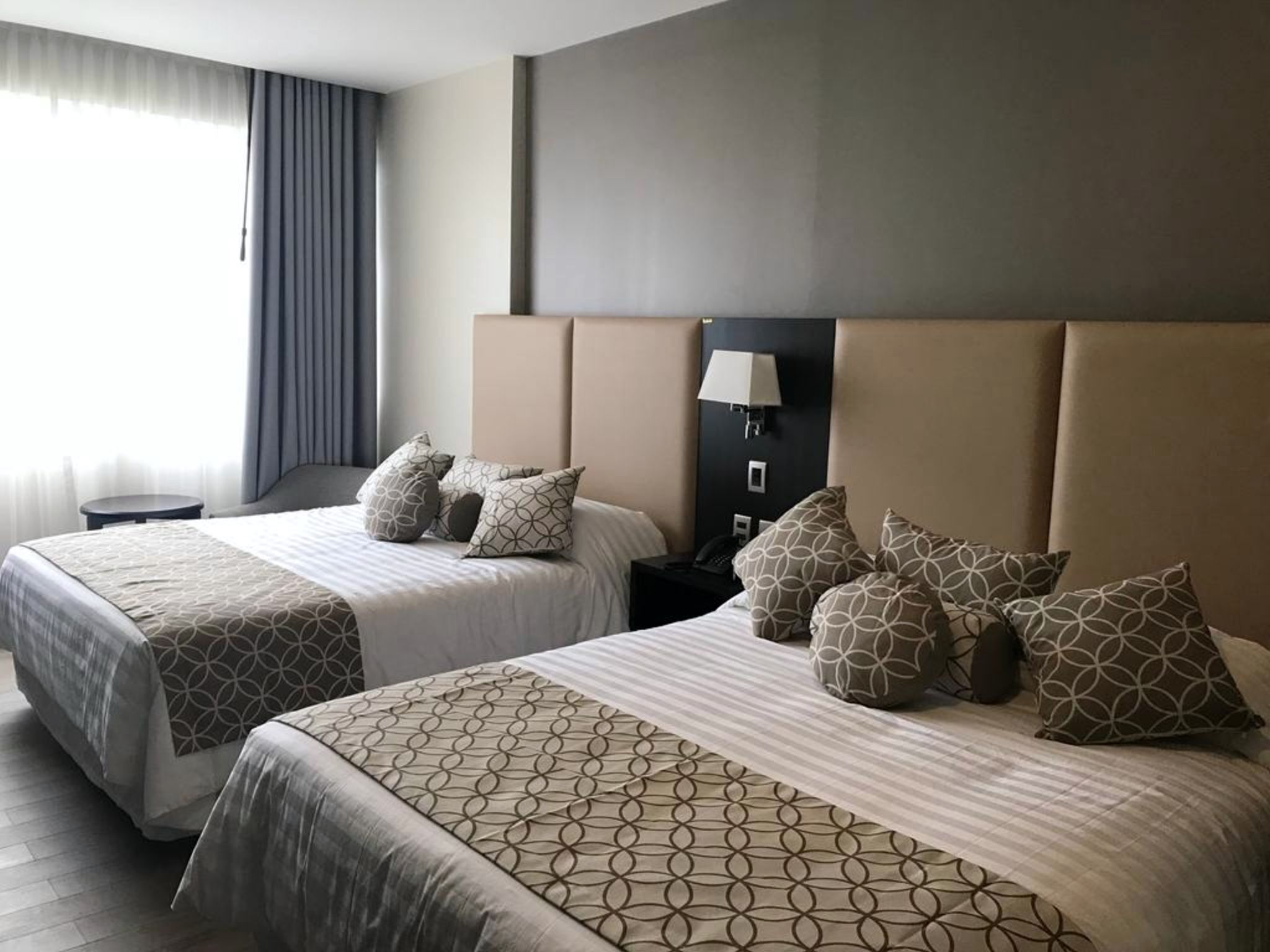 Habitación de hotel con dos camas, una mesita de noche, una lámpara y una ventana con cortinas.