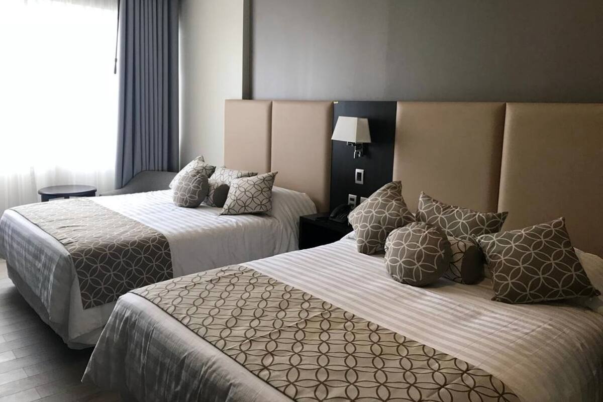 A hotel room with two beds, each with patterned bedding and pillows, a bedside table, a lamp, and a view of the outdoors.