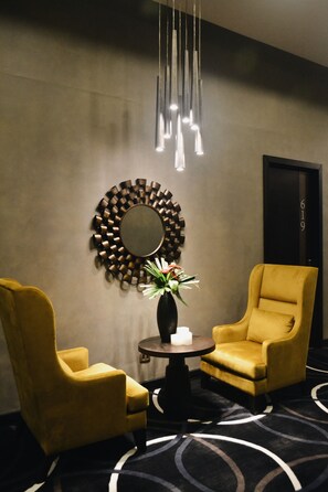 A modern room with two yellow armchairs, a round table with a vase of flowers, a wall-mounted mirror, and a hanging light fixture.