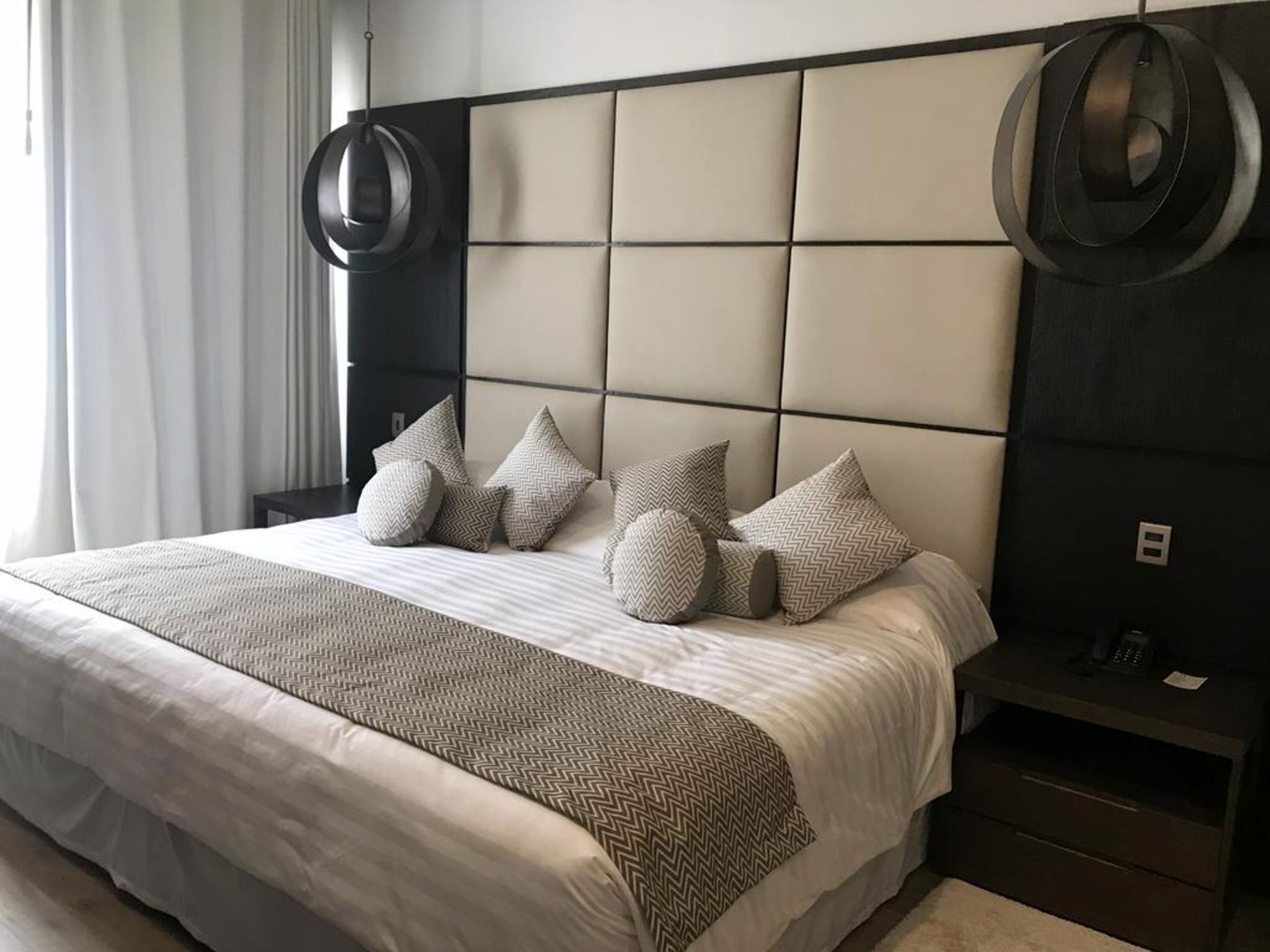Una habitación de hotel moderna con una cama grande, un cabecero con un diseño geométrico y una mesita de noche con un teléfono y una lámpara pequeña.