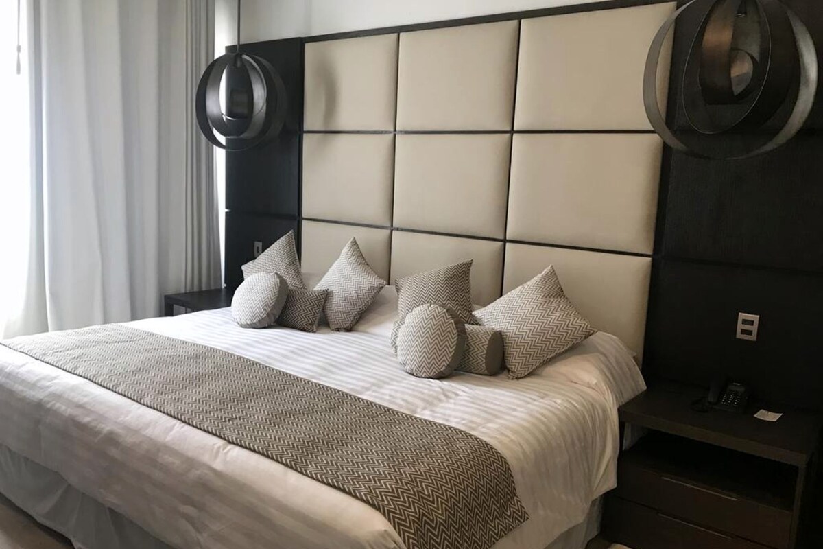 Una habitación de hotel moderna con una cama grande, un cabecero con diseño geométrico y una mesita de noche con un teléfono y una lámpara.