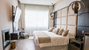 Habitación de hotel moderna con una cama grande, una mesita de noche, un escritorio, una silla, un televisor y un ventanal con cortinas.