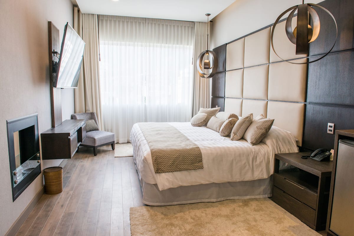 A modern hotel room with a large bed, a bedside table, a desk, a chair, a TV, and a large window with curtains.