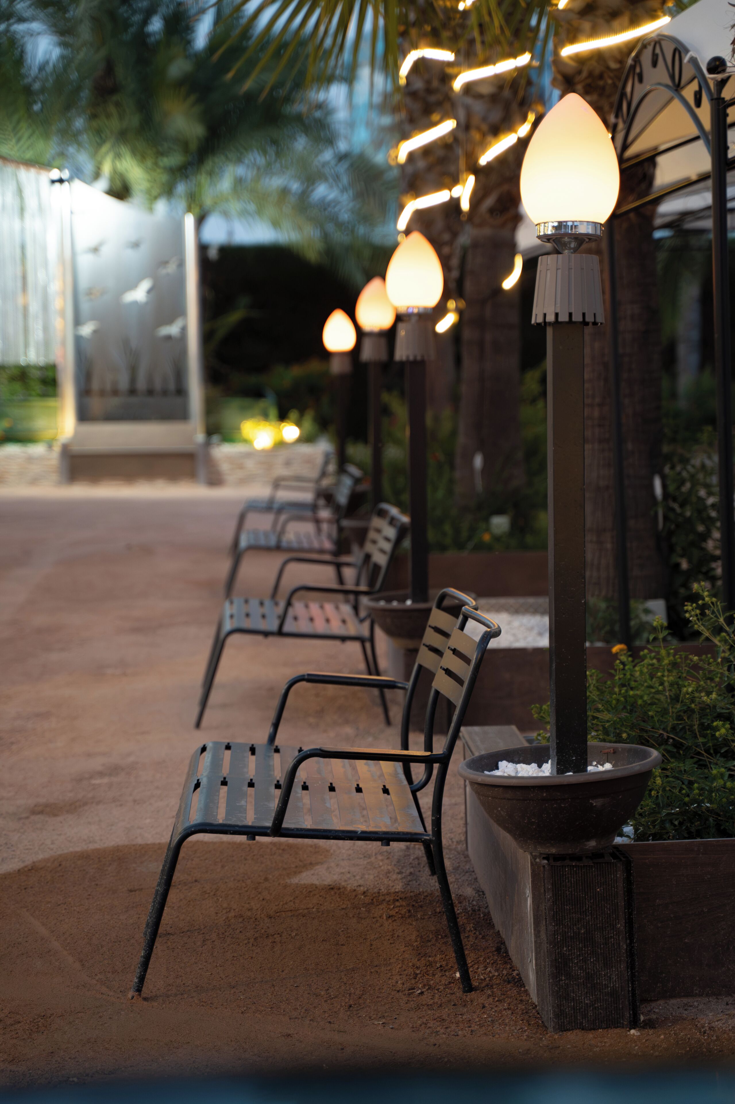 Mobiliario exterior con sillas de metal y una jardinera, bajo un farol.