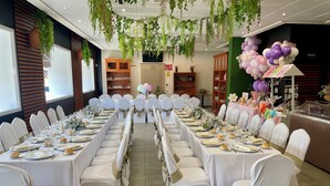 Un salón de banquetes con largas mesas dispuestas para un evento formal, rodeado de sillas blancas con cintas doradas, y un techo decorado con plantas colgantes.