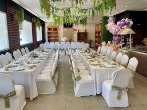 Un salón de banquetes con largas mesas dispuestas para un evento formal, rodeado de sillas blancas con cintas doradas, y un techo decorado con plantas colgantes.