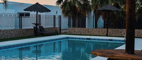 Piscina con sillones de descanso y sombrillas, rodeada de un muro de piedra y palmeras.