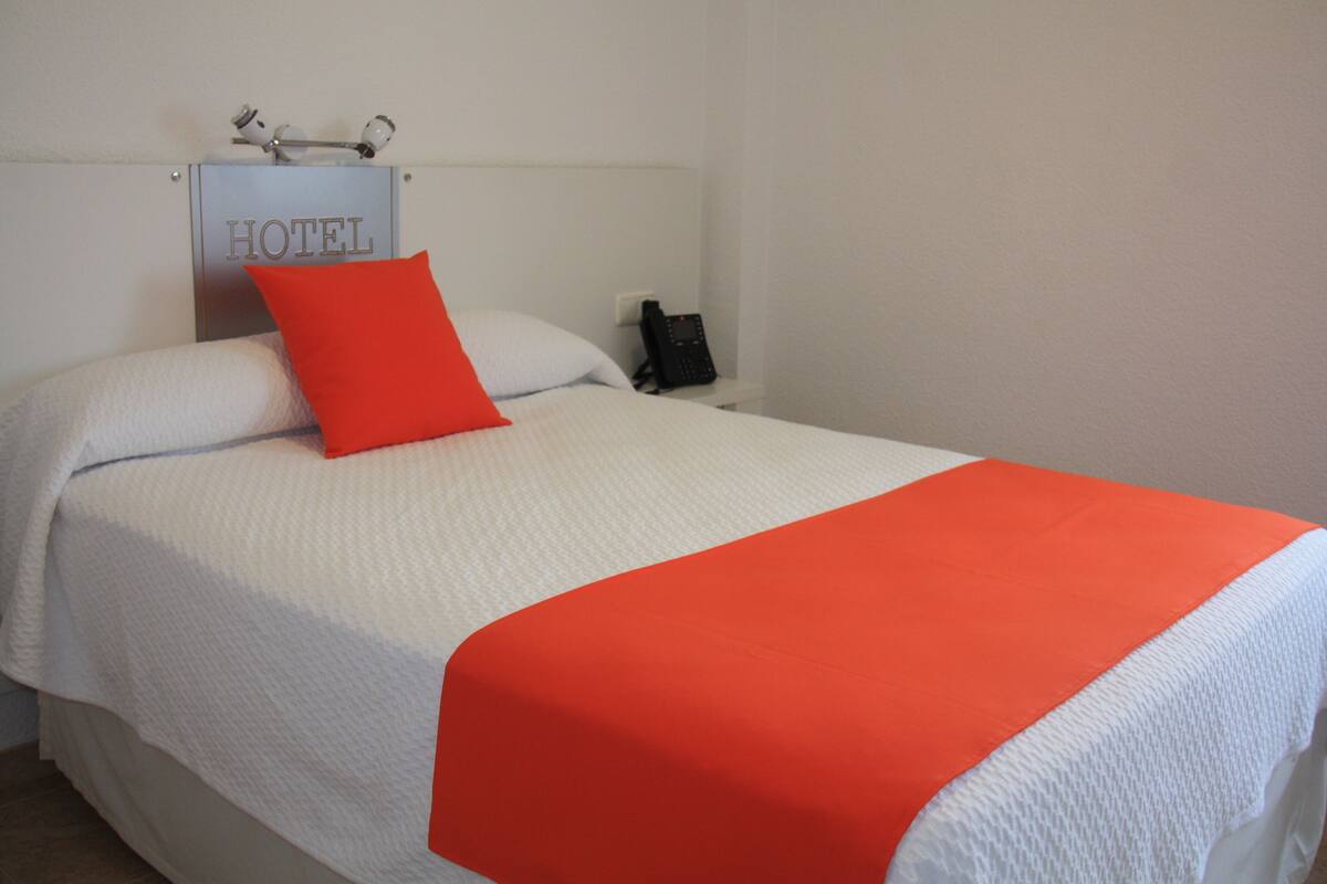 Una habitación de hotel con una cama, un almohadón rojo, una colcha blanca, un cabecero blanco y un teléfono en la mesita de noche.