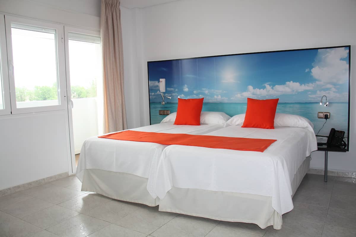 Cama doble con ropa de cama blanca y almohadas rojas, una mesita de noche con teléfono y un ventanal con cortinas.