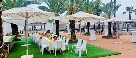 An outdoor dining area with white tables and chairs, surrounded by palm trees and umbrellas.