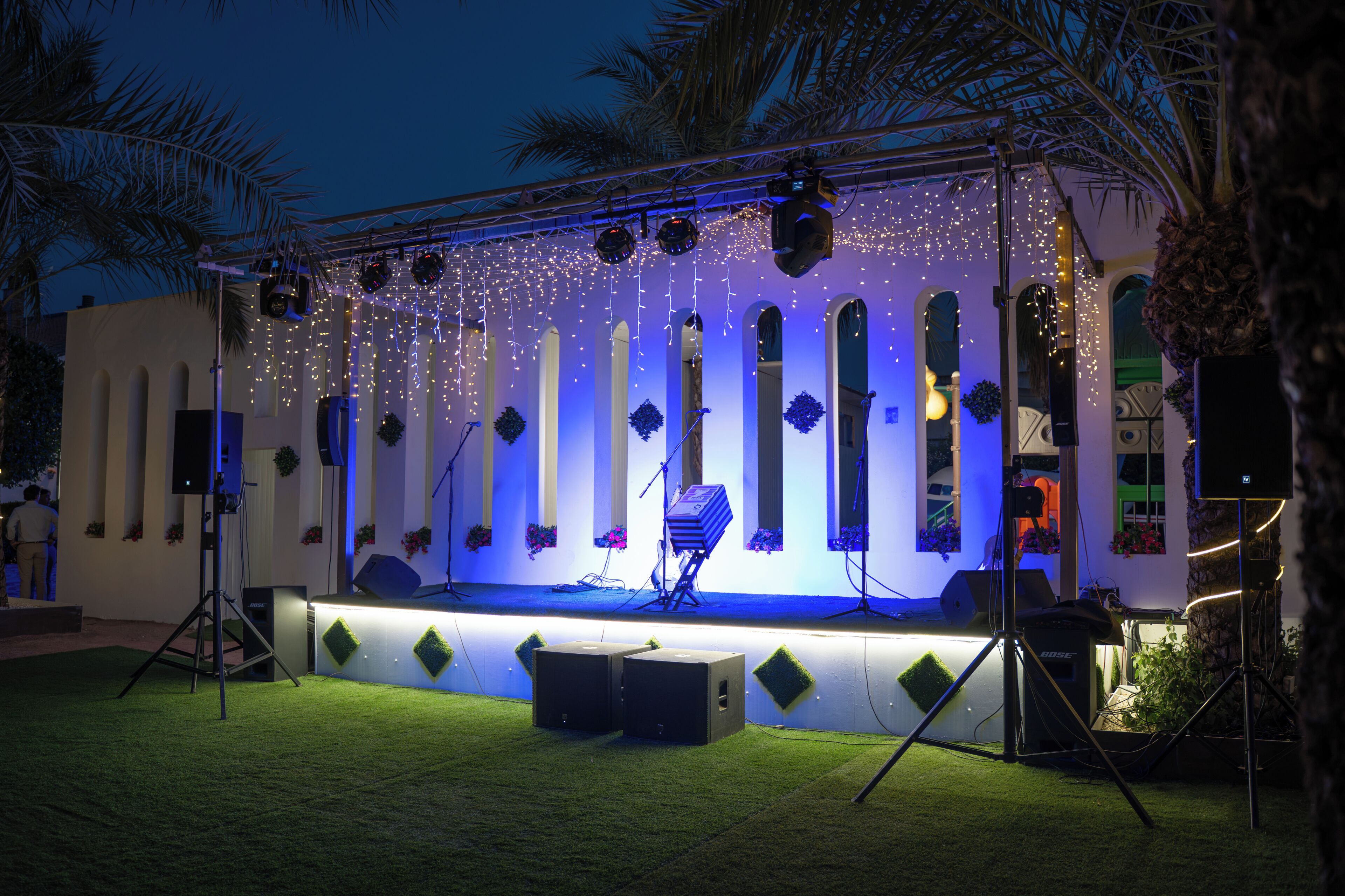 Un escenario al aire libre con un fondo de arcos y luces colgantes, rodeado de follaje y palmeras.