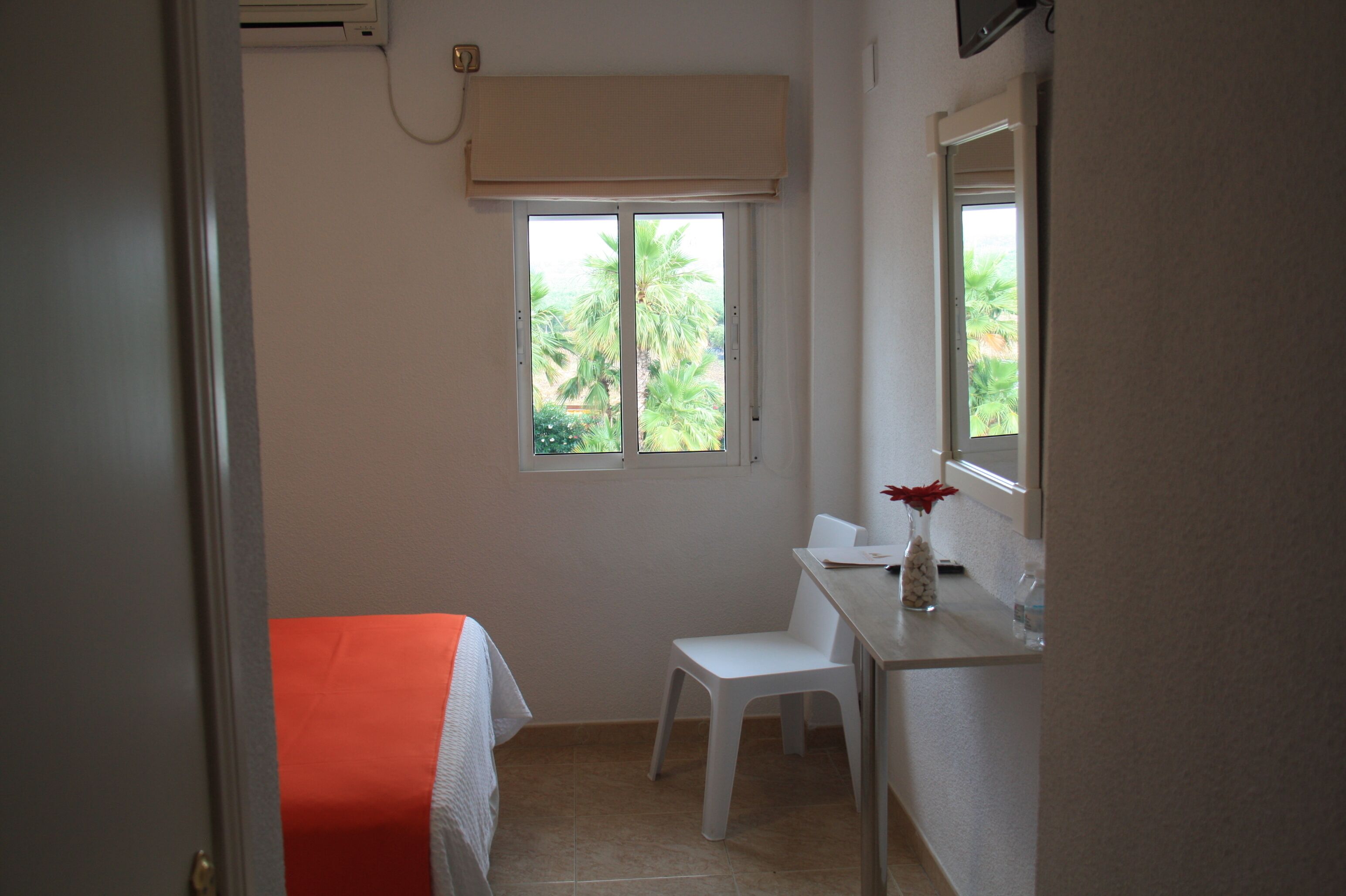 A small, clean room with a bed, a white chair, a desk, and a window with a view of palm trees.