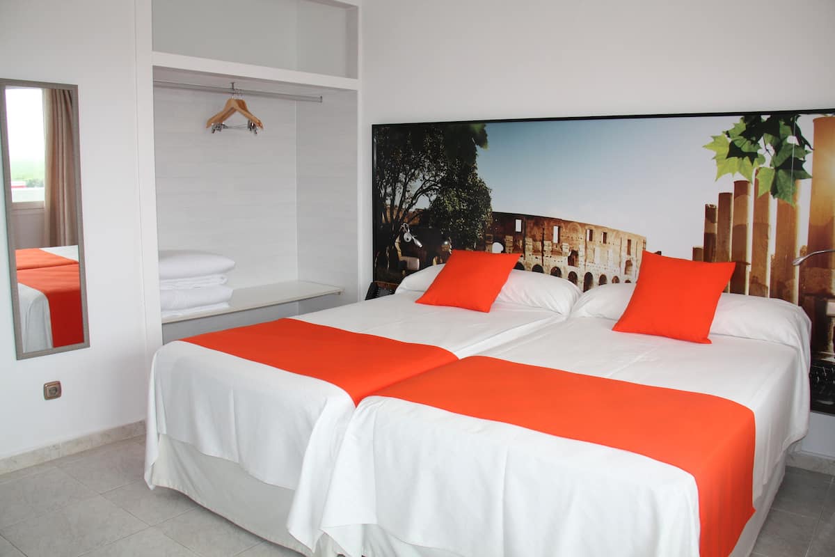 Habitación de hotel con dos camas, ropa blanca, detalles en naranja, un ventanal grande y un mural de una estructura antigua.