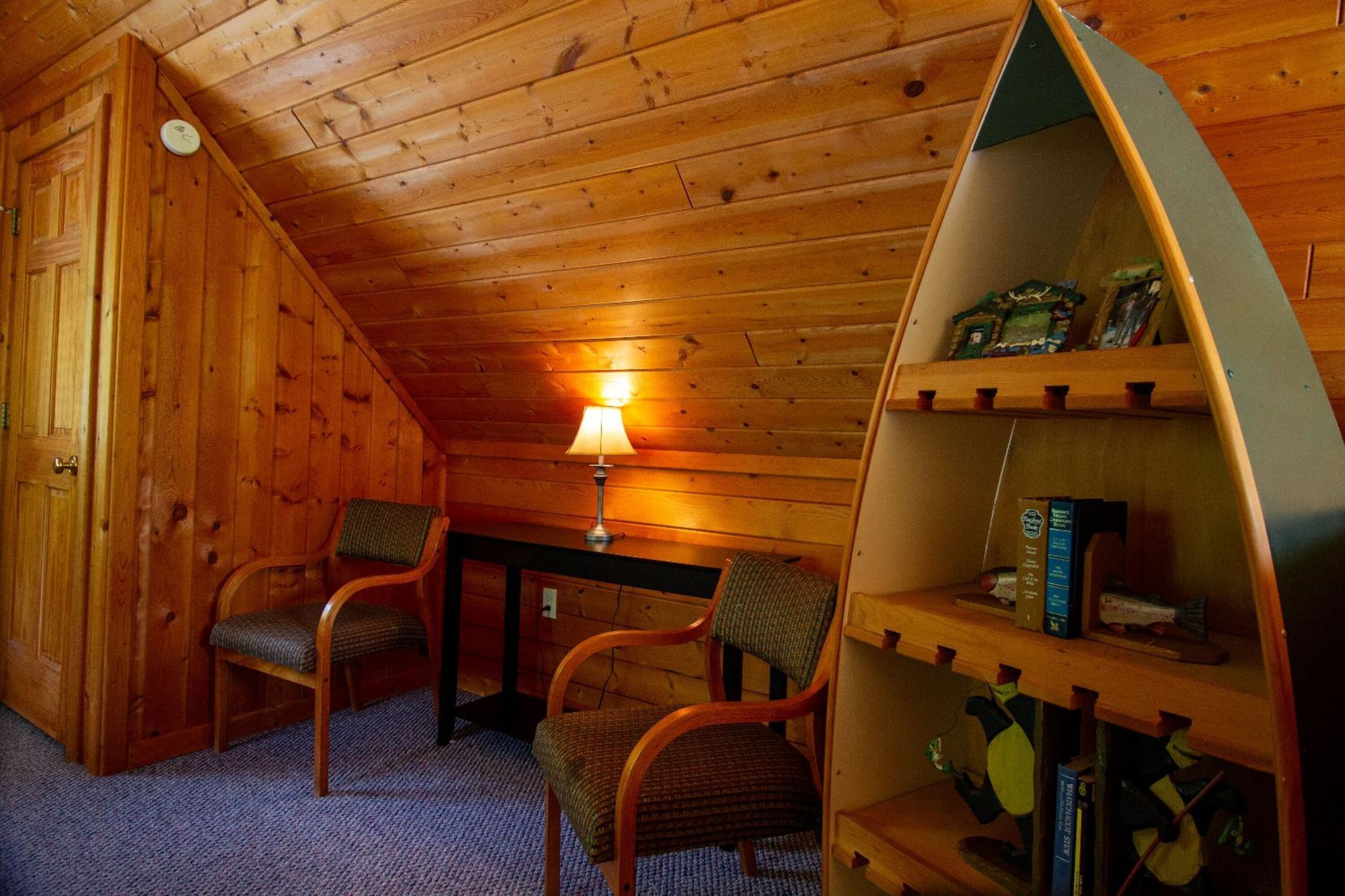 Una habitación acogedora con paredes y techo de madera, que incluye dos sillones, una mesa con lámpara y una estantería.