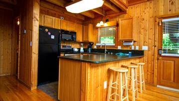 A wooden cabin kitchen with a central island, bar stools, a refrigerator, microwave, and a window with blinds.