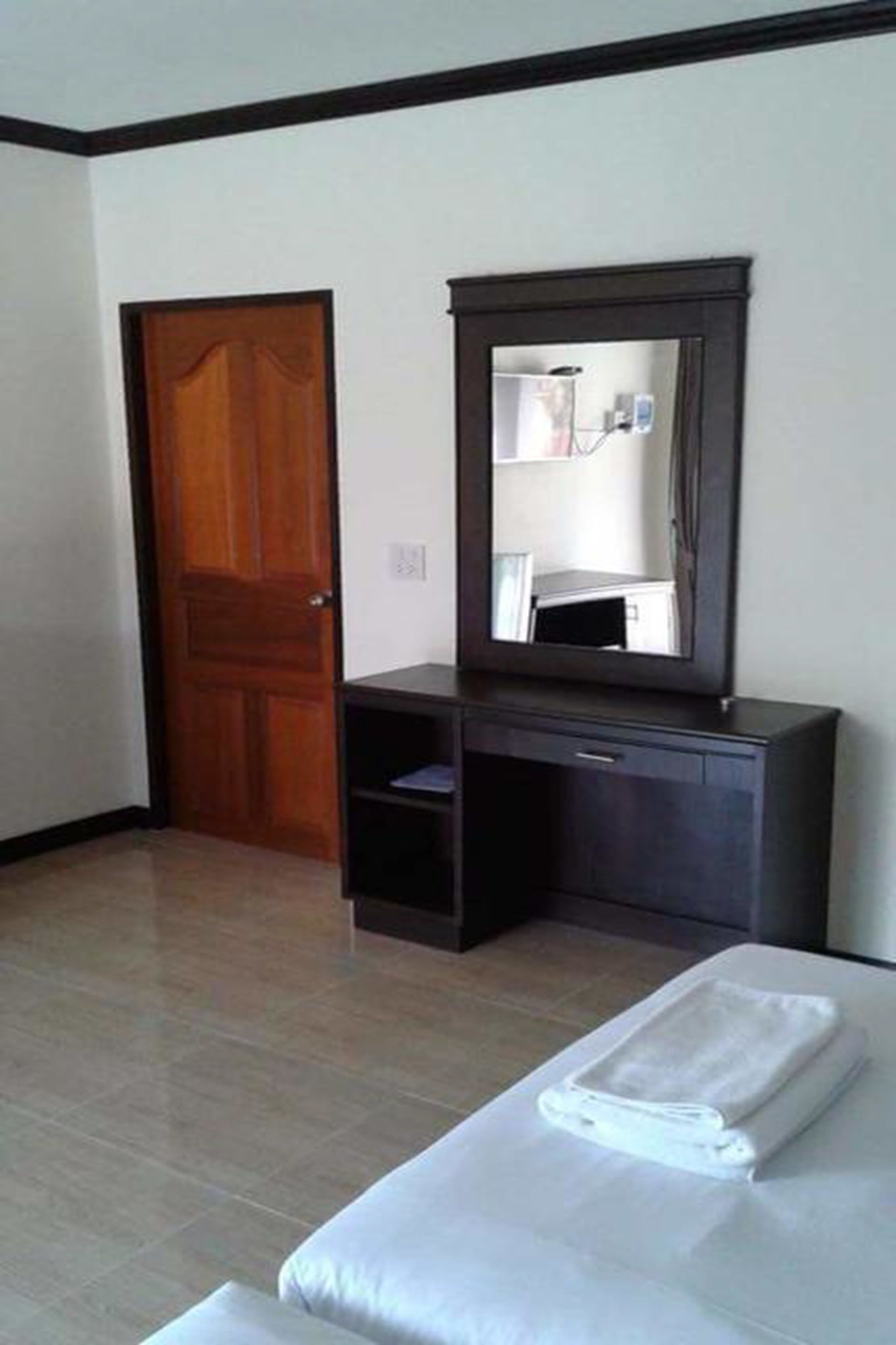 A hotel room with a wooden door, a dark-colored dresser with a mirror, and a neatly made bed with a white blanket.