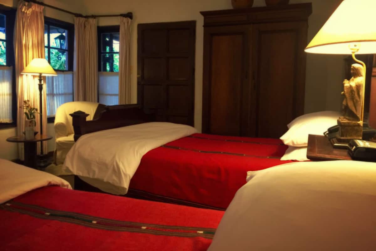 Quarto de hotel com duas camas, um guarda-roupa de madeira, uma luminária e uma janela com cortinas.