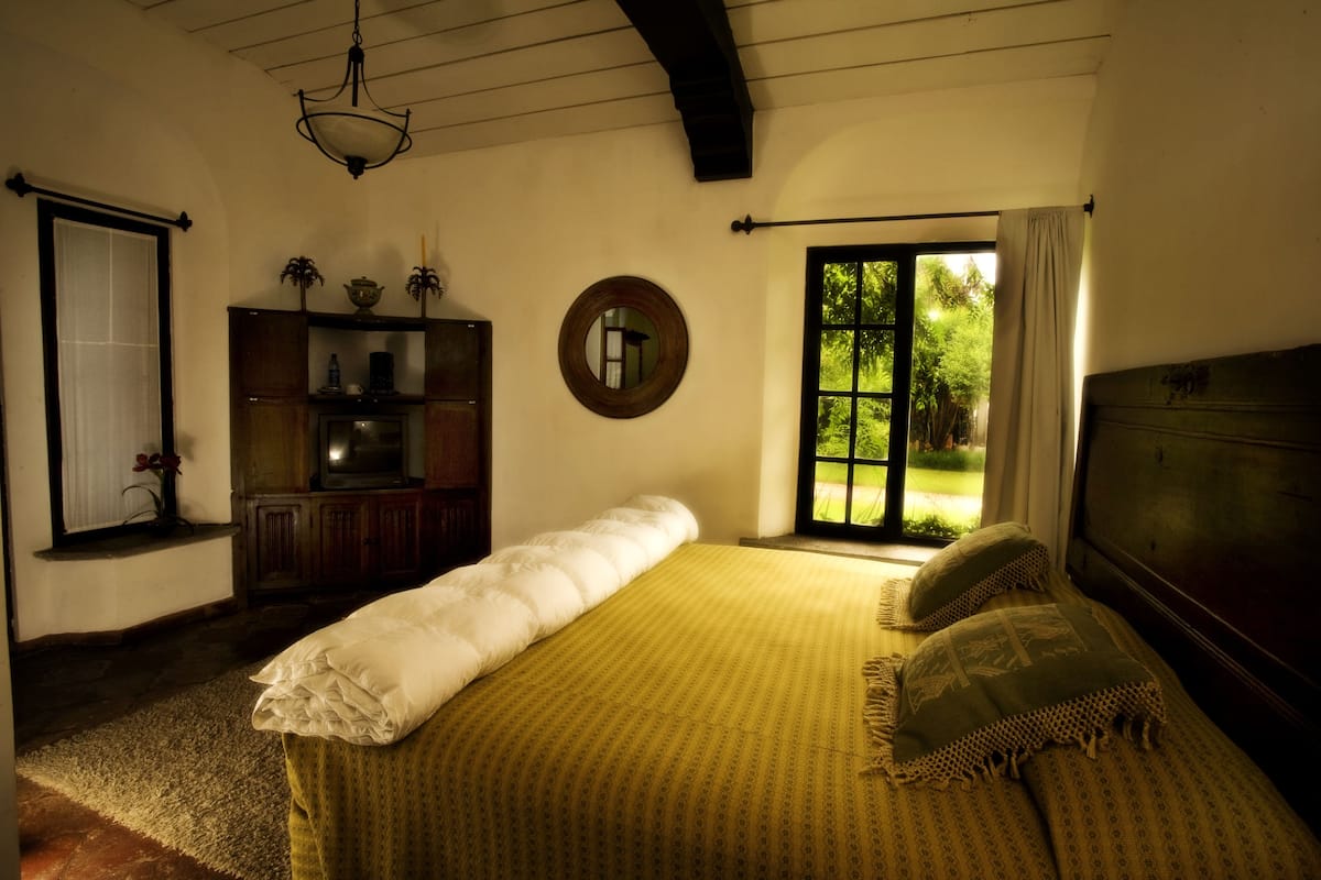 Quarto com cama grande, armário de madeira, espelho e janela com vista para o verde.