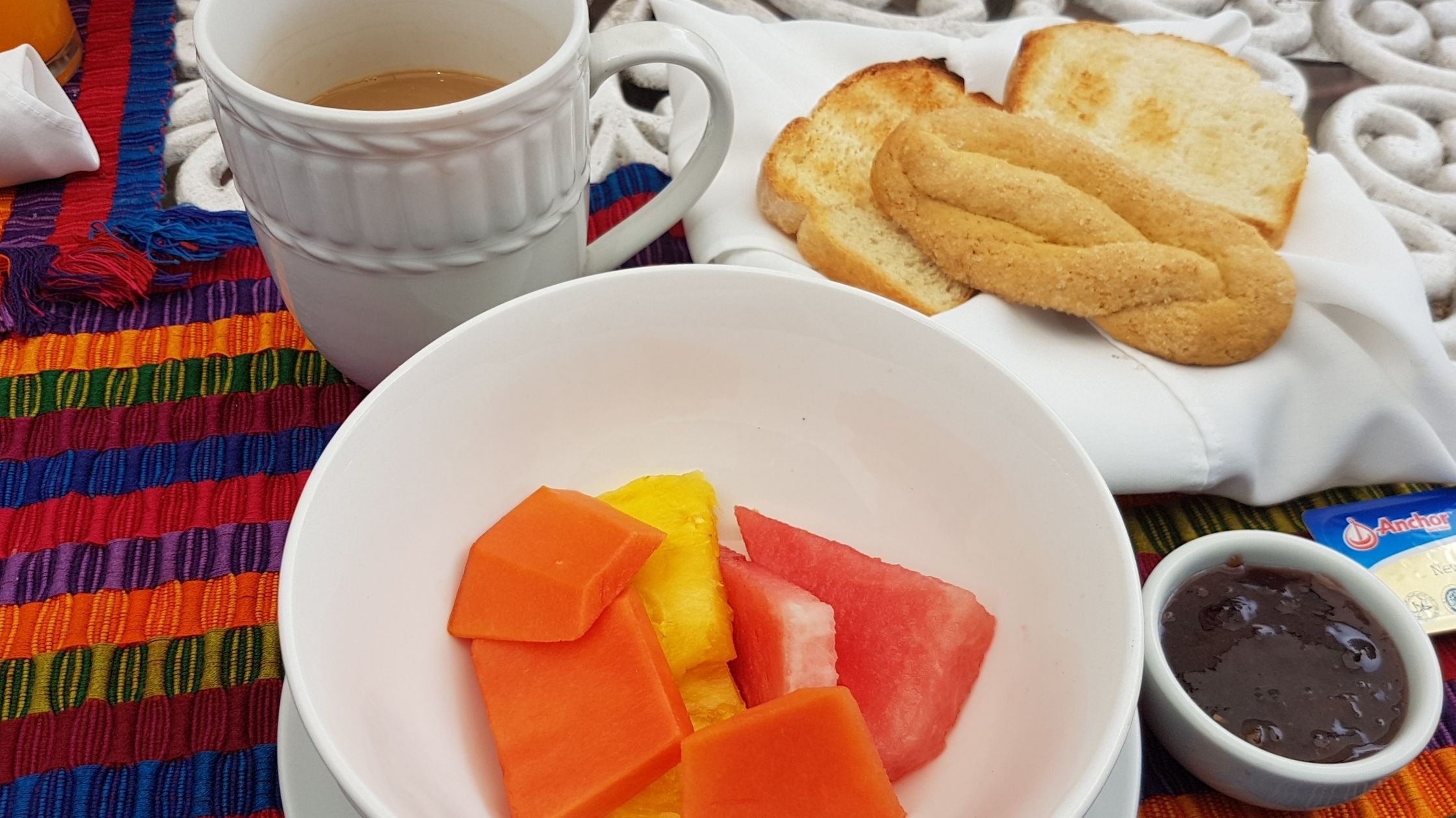 Une table mise pour le petit-déjeuner avec une tasse de café, des toasts, un bol de fruits et un petit pot de pâte à tartiner au chocolat.