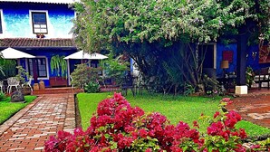 A blue building with a tiled roof, a brick pathway, and a garden with red flowers. - Casa Capuchinas (Antigua Guatemala)