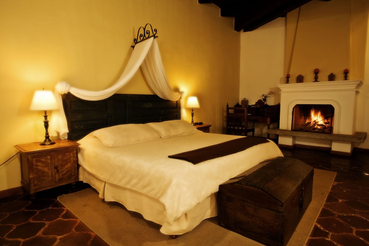 Quarto com uma cama grande, mesa de cabeceira com luminária, um banquinho e lareira.