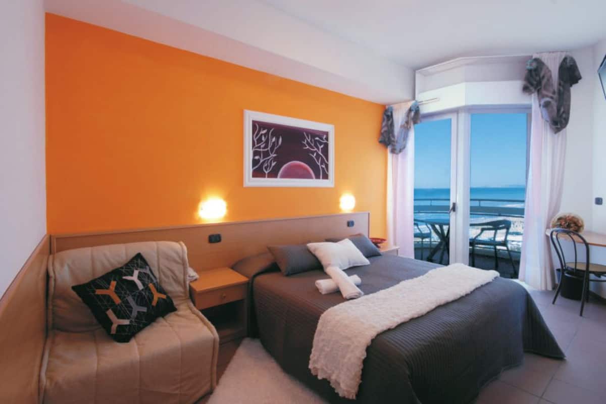 Une chambre d’hôtel avec un mur orange, un lit recouvert d’une couverture blanche, une table de chevet avec une lampe, et une vue sur l’océan par une porte coulissante.