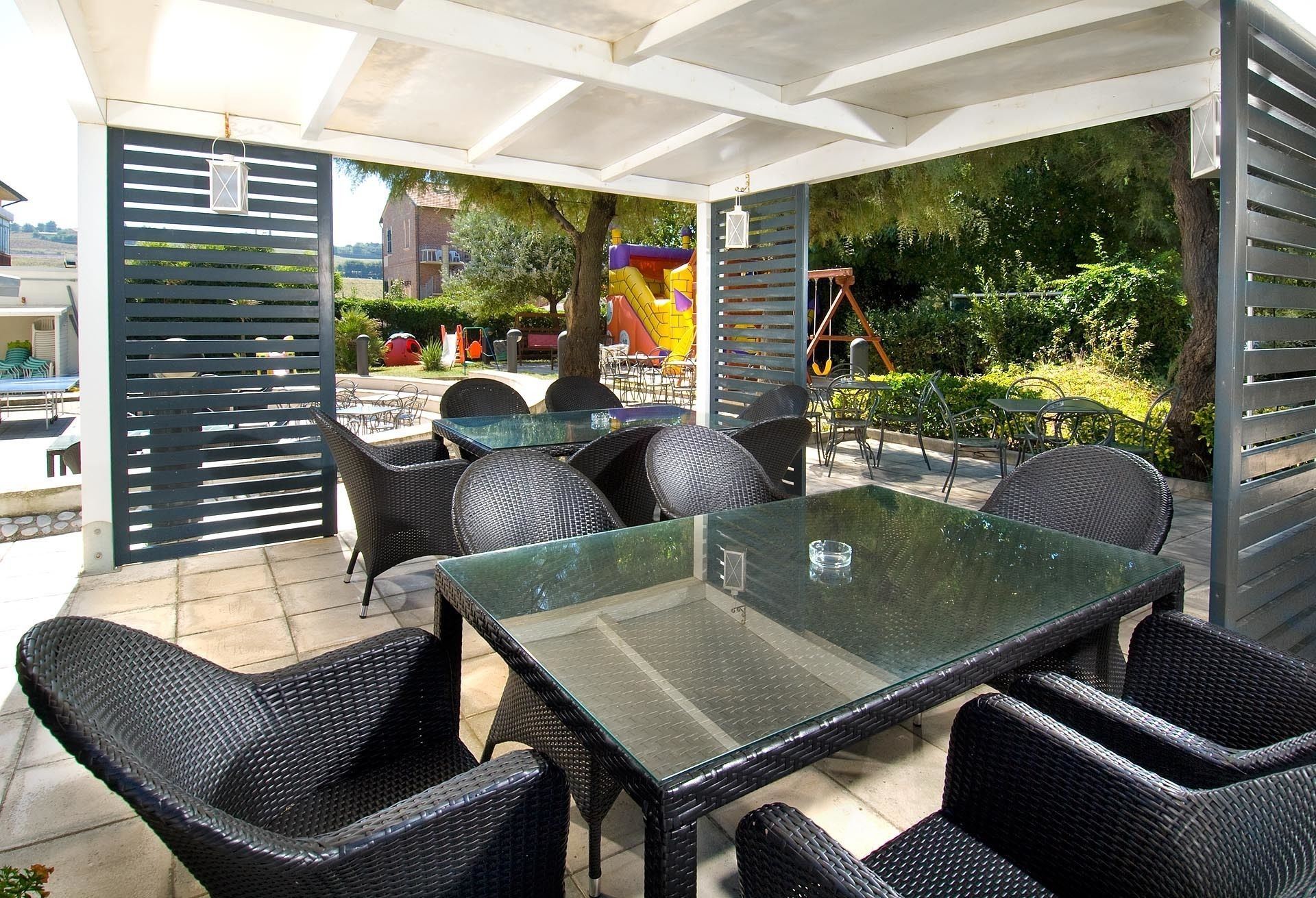 An outdoor patio with a glass-top table and wicker chairs, surrounded by a white pergola structure.