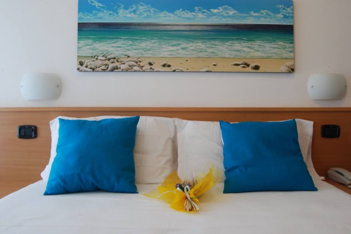 Une chambre d’hôtel avec un lit, deux oreillers bleus, un oreiller blanc, un bouquet de fleurs jaunes, un tableau représentant une plage et un téléphone sur la table de chevet.