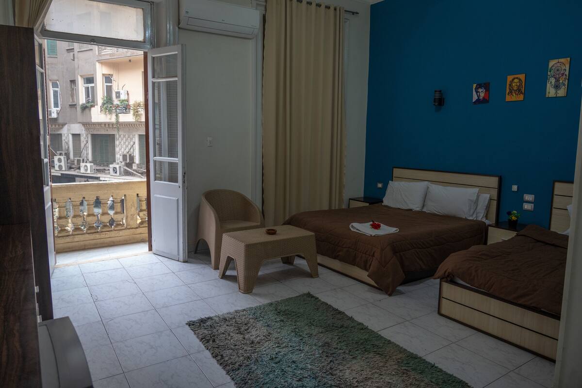 Ein Hotelzimmer mit einem Bett, einem Sessel, einem kleinen Tisch, einer blauen Wand mit Kunstwerken und einem Balkon mit Blick ins Freie.