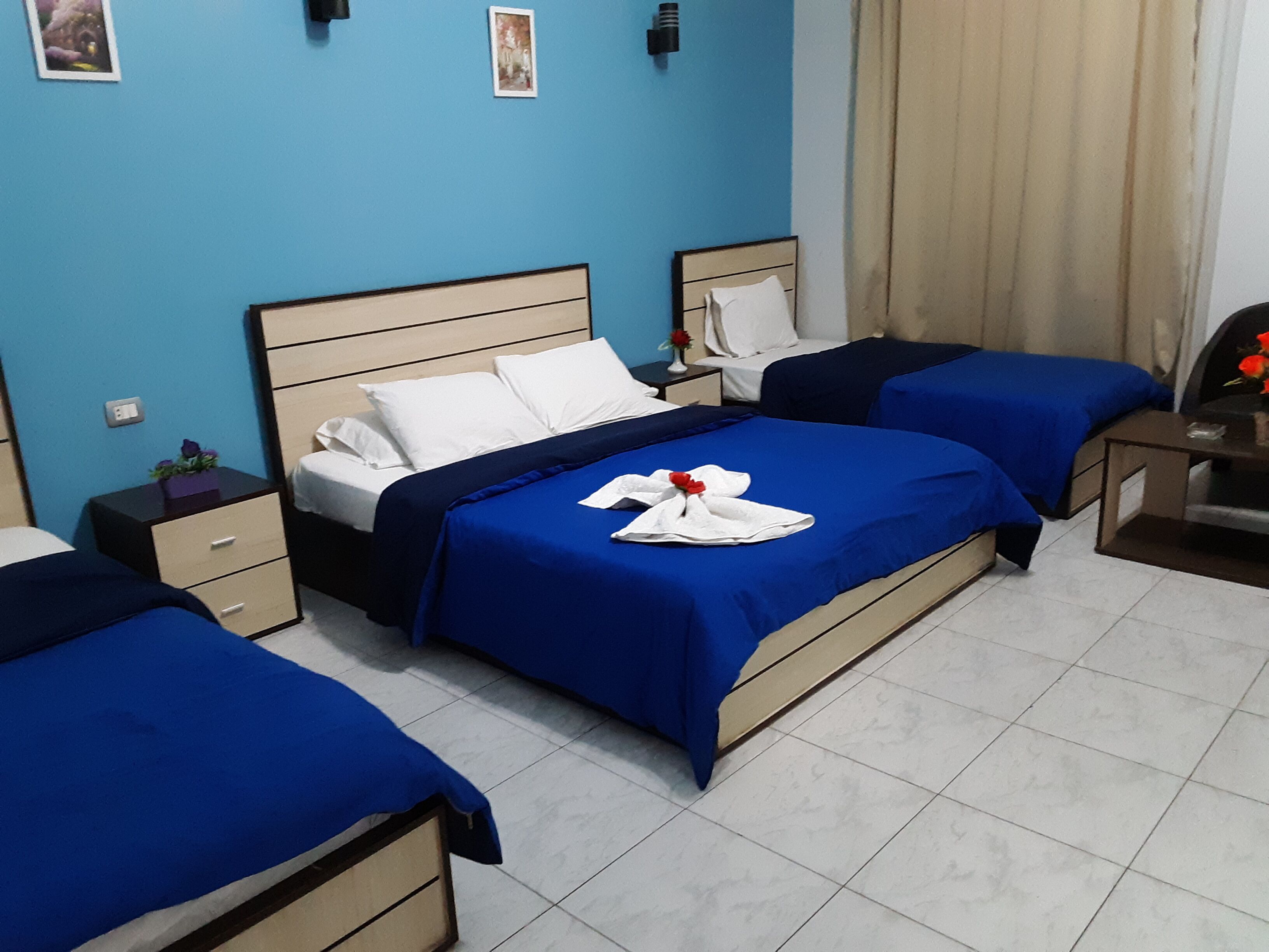Habitación de hotel con dos camas, cada una con cubrecama azul y almohadas blancas. Hay una mesita de noche con una lámpara y una pequeña mesa con un florero.