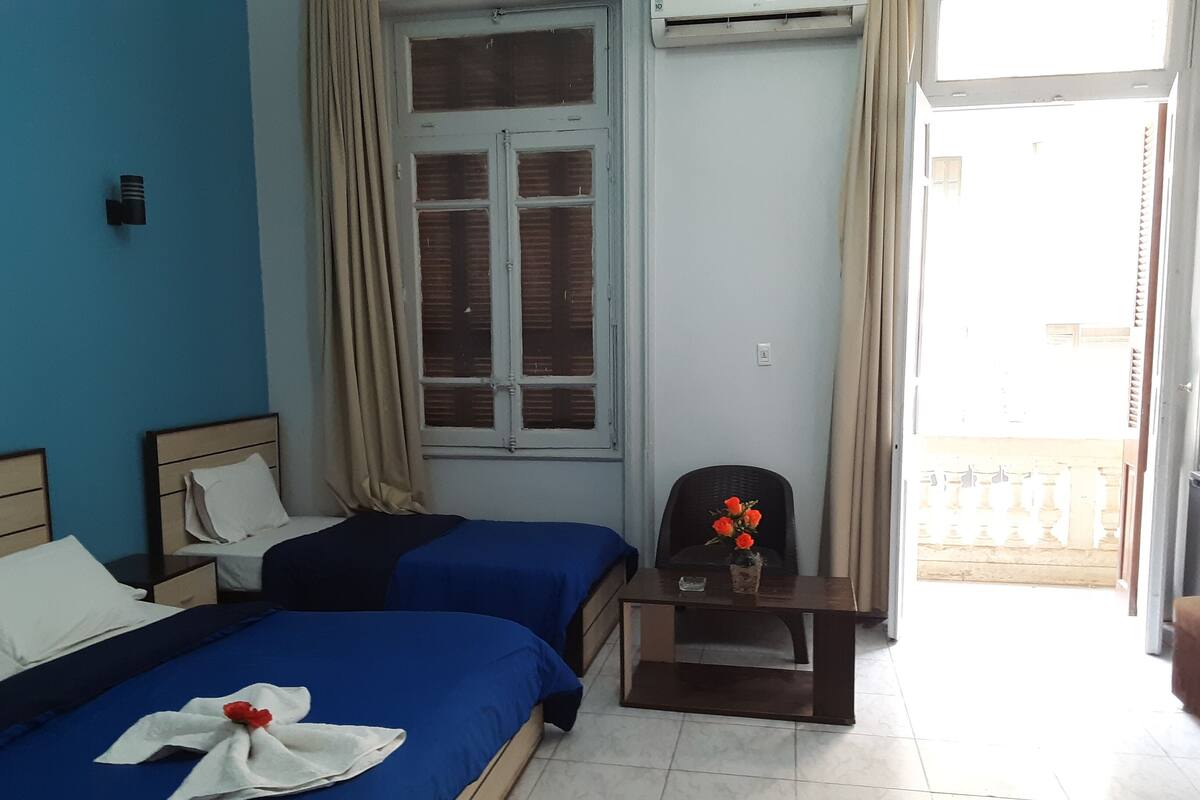 Ein Hotelzimmer mit zwei Betten, einem kleinen Tisch mit Blumenvase, einem Sessel, einem Fenster mit Vorhängen und einer Tür zu einem Außenbereich.