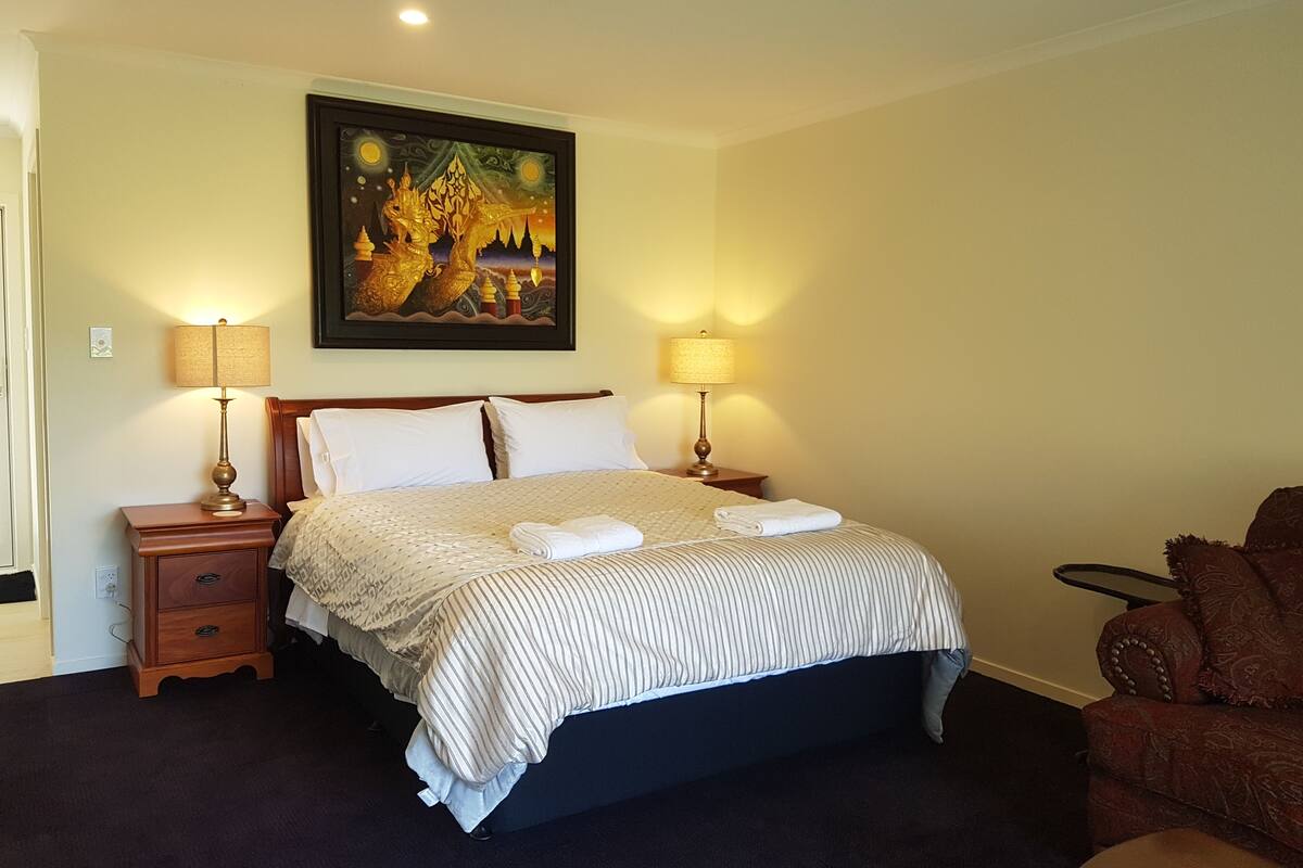 A hotel room with a large bed, two bedside lamps, a nightstand, a chair, and a painting on the wall.
