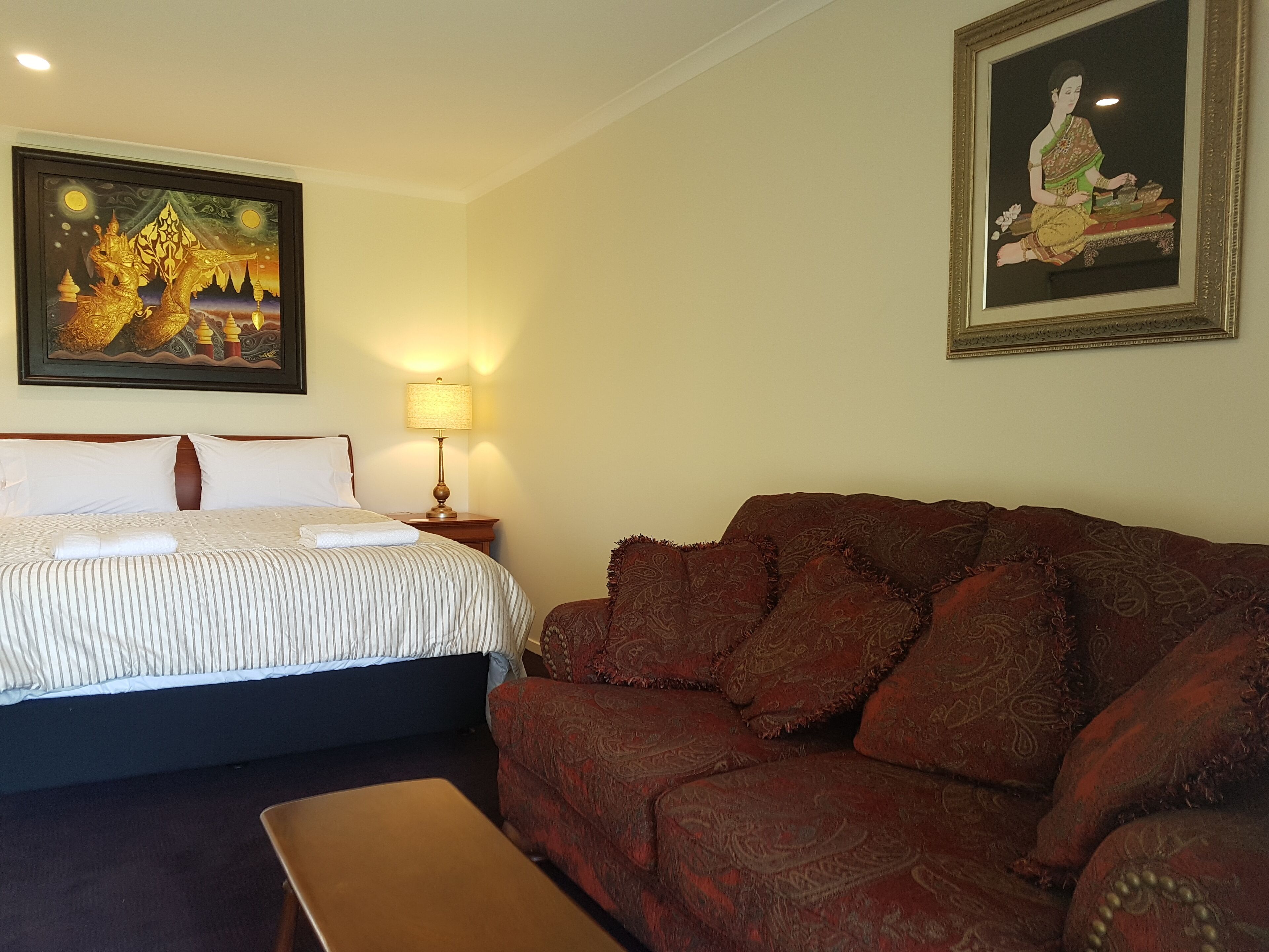 Una camera d'albergo con un letto, un divano rosso con motivo decorativo, un comodino con una lampada e due opere d'arte incorniciate alle pareti.