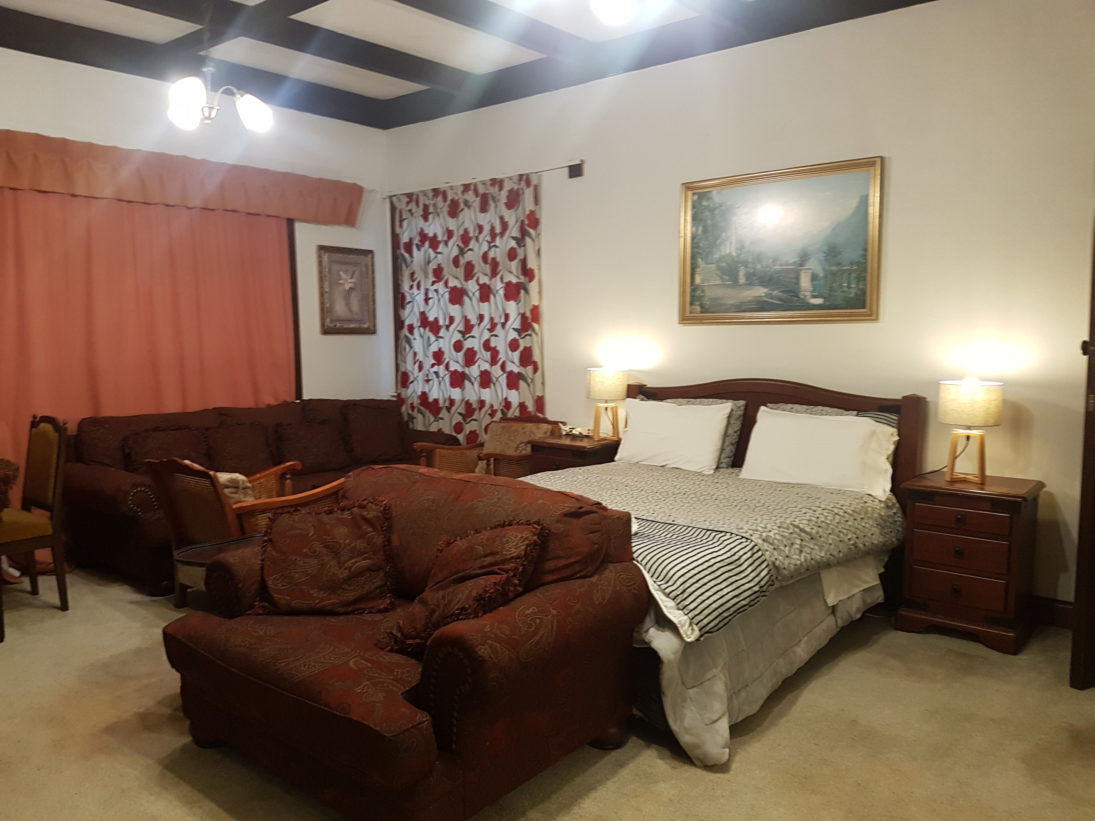 Un dormitorio con cama, dos mesitas de noche, cabecera de madera, un cuadro, cortina floral y un sillón marrón.