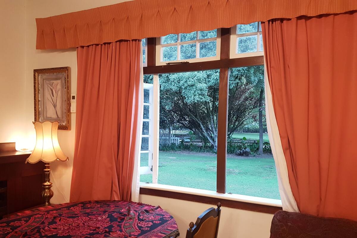 A room with a large window, a table with a patterned tablecloth, a chair, and a lamp.