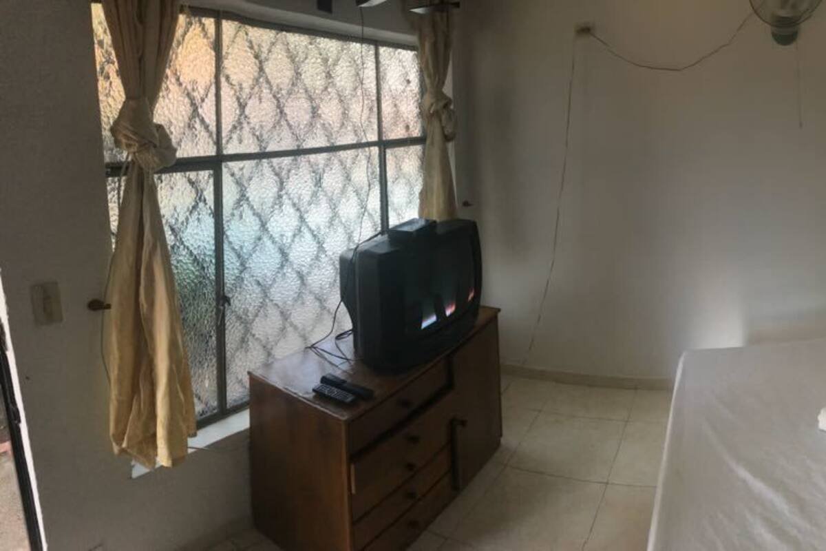 Una habitación con una cama, un televisor sobre un tocador de madera y una ventana con cortinas.
