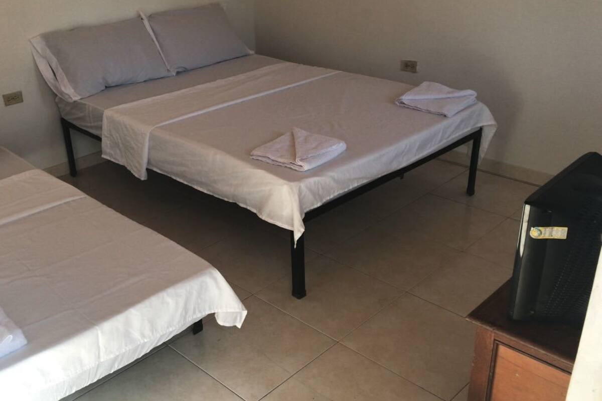 Una habitación con dos camas individuales, cada una con cubrecama y almohadas blancas, y un escritorio pequeño con un objeto negro encima.