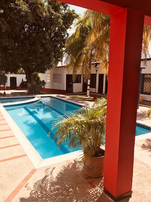 A swimming pool with a red column in the foreground, surrounded by palm trees and a building with a white facade. - Quinta Real Amaranta (Ricaurte)