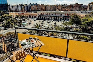 Ein Balkon mit Tisch für Zwei, Blick auf einen Stadtplatz mit Gebäuden und einem Brunnen.