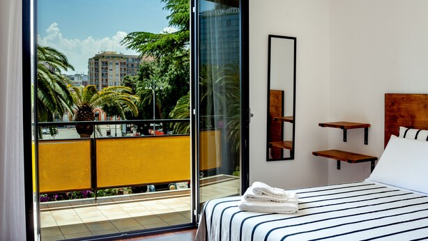 A hotel room with a large sliding glass door leading to a balcony with a view of palm trees and buildings. There is a bed with striped bedding and a wooden headboard. - Travel B&B (Bari)