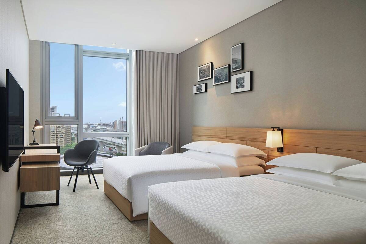 Een hotelkamer met twee bedden, een bureau, een stoel, een televisie en een raam met uitzicht op de stad.