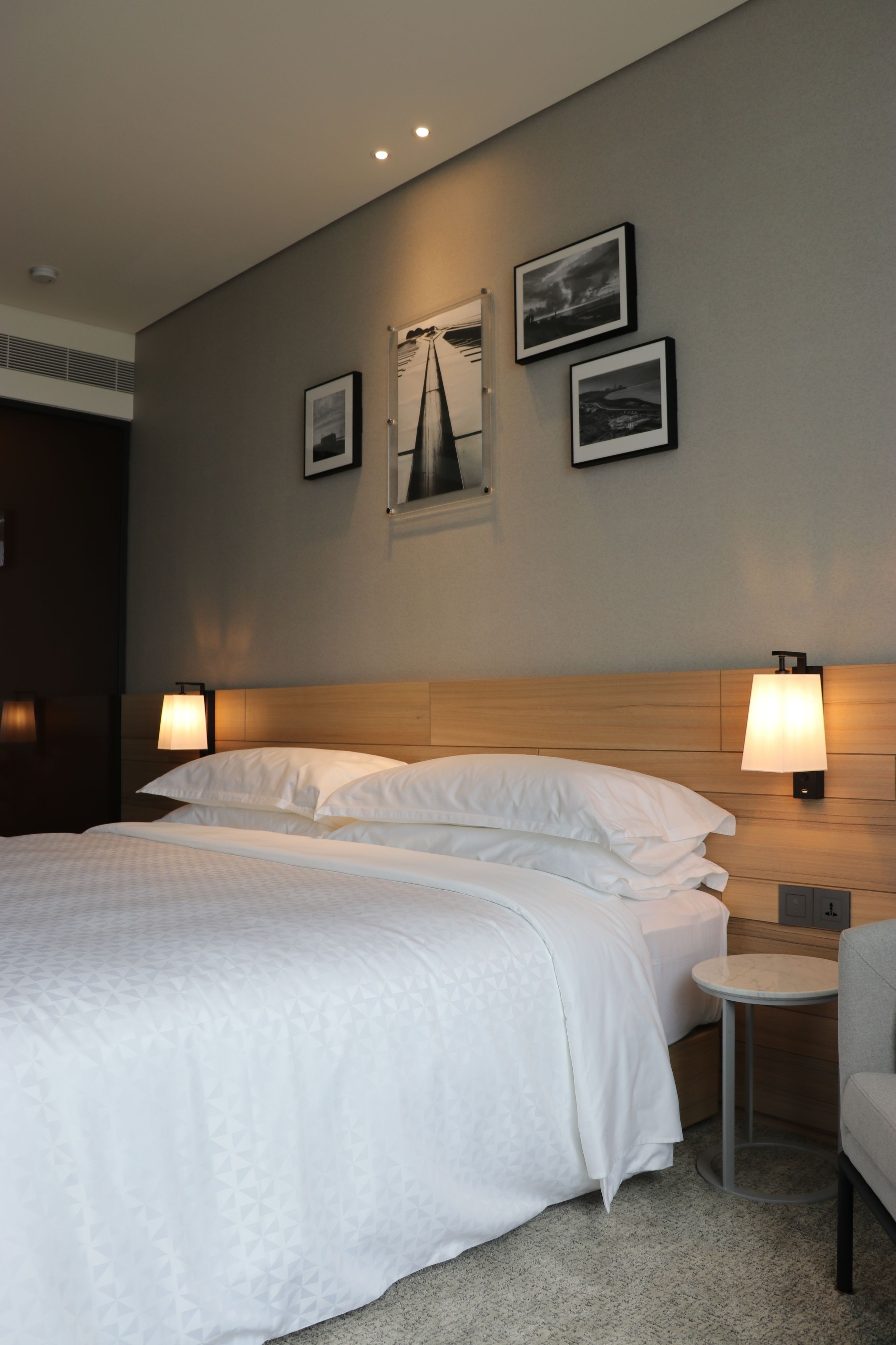 Habitación de hotel con una cama grande, dos lámparas de noche, una mesita redonda y una silla. En la pared sobre la cama hay cuadros enmarcados.