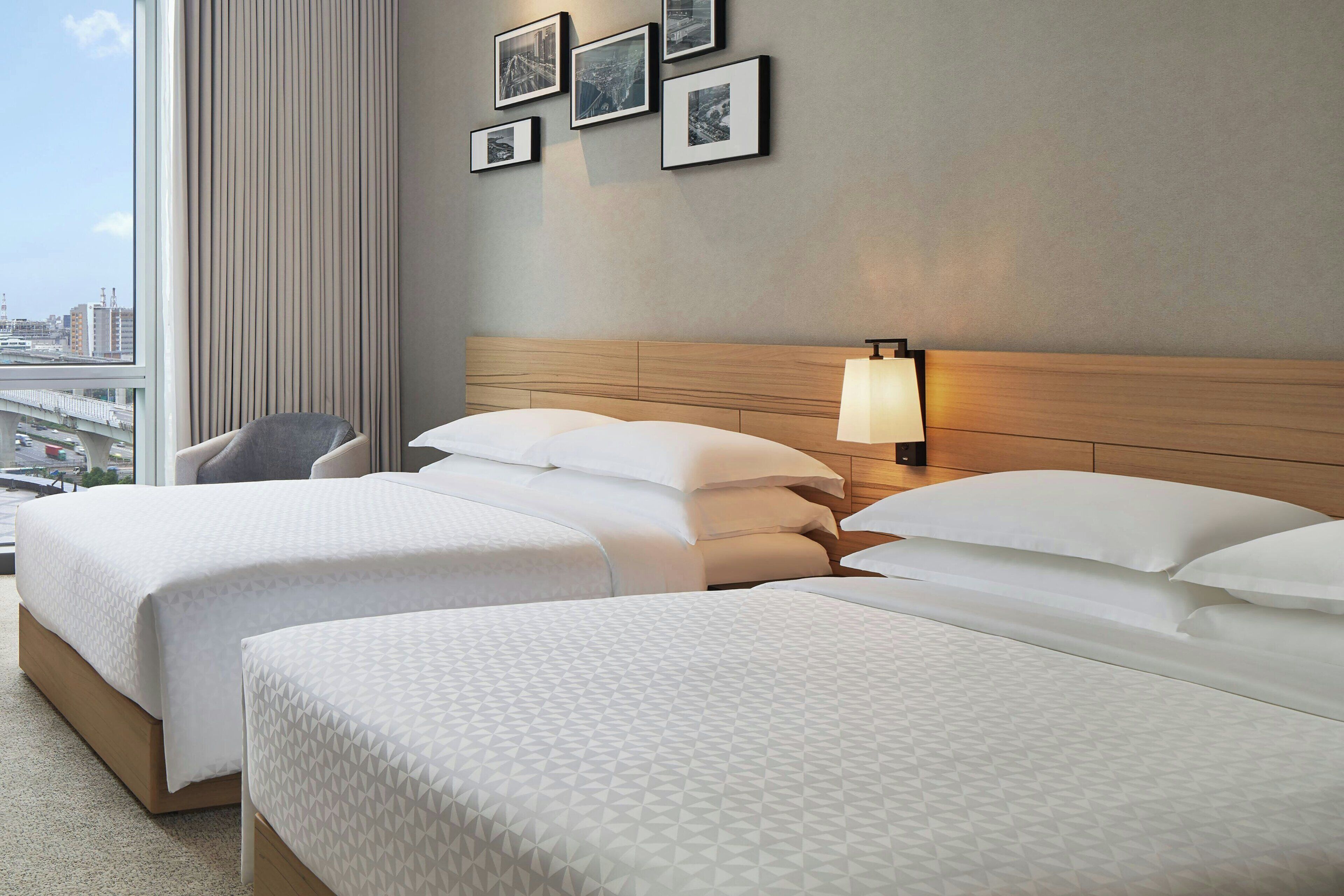 Een hotelkamer met twee bedden, een stoel, uitzicht op de stad en ingelijste foto's aan de muur.
