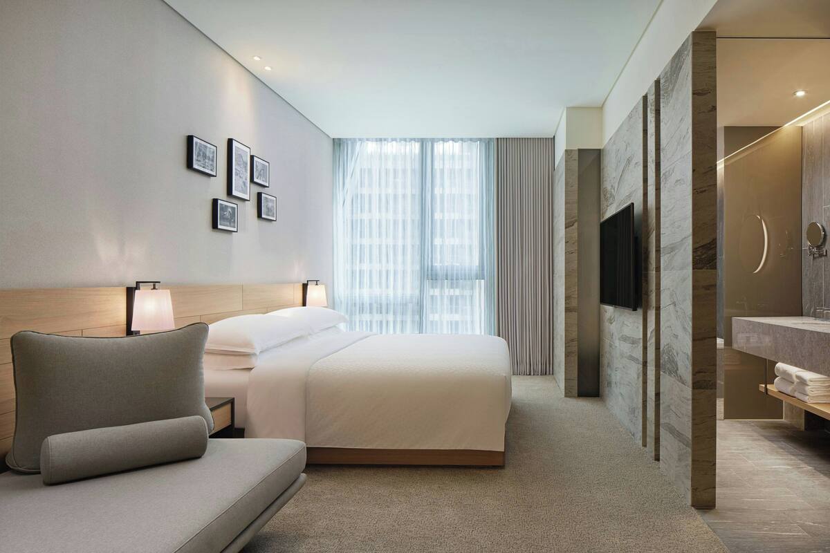 Een moderne hotelkamer met een groot bed, een bank, een televisie en een badkamer met spiegel en wastafel.