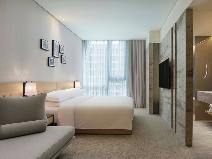 A modern hotel room with a large bed, a sofa, a TV, and a bathroom with a mirror and sink.