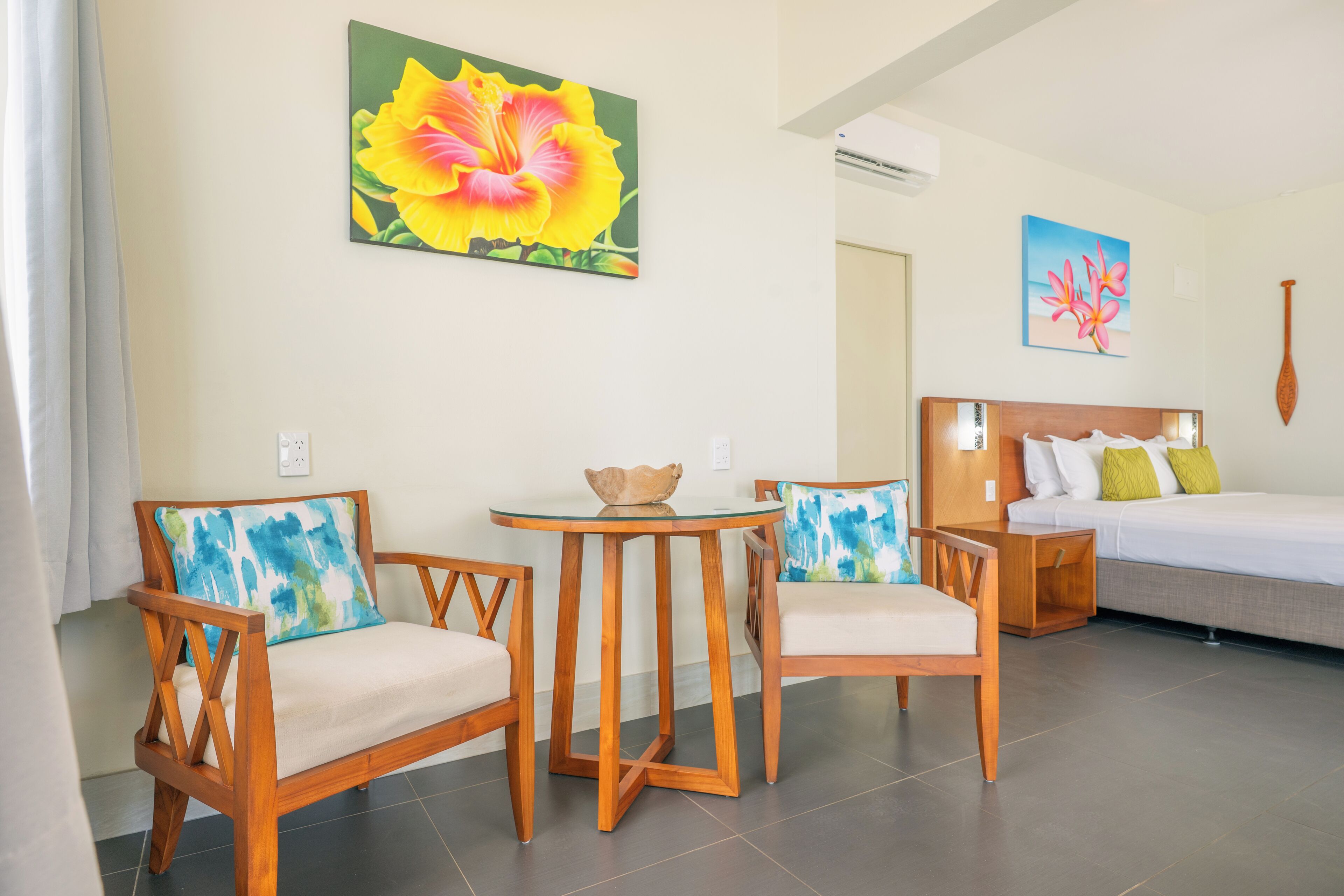 Habitación de hotel con una cama, dos sillas de madera, una mesita redonda y dos cuadros colgados en la pared.